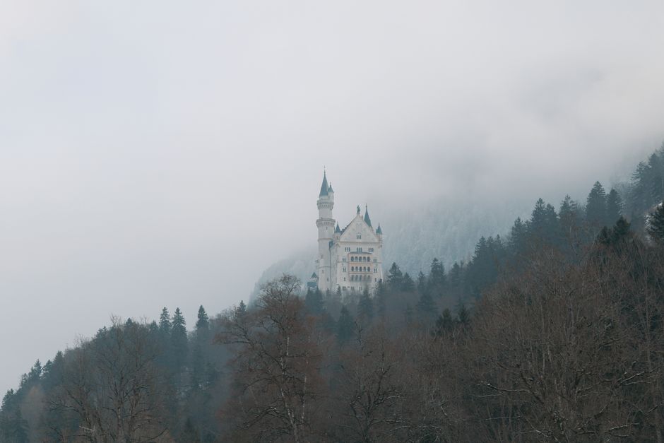 Dramatic view of Neuschwanstein Castle surrounded by winter fog and forested hills in Schwangau, Bavaria.
